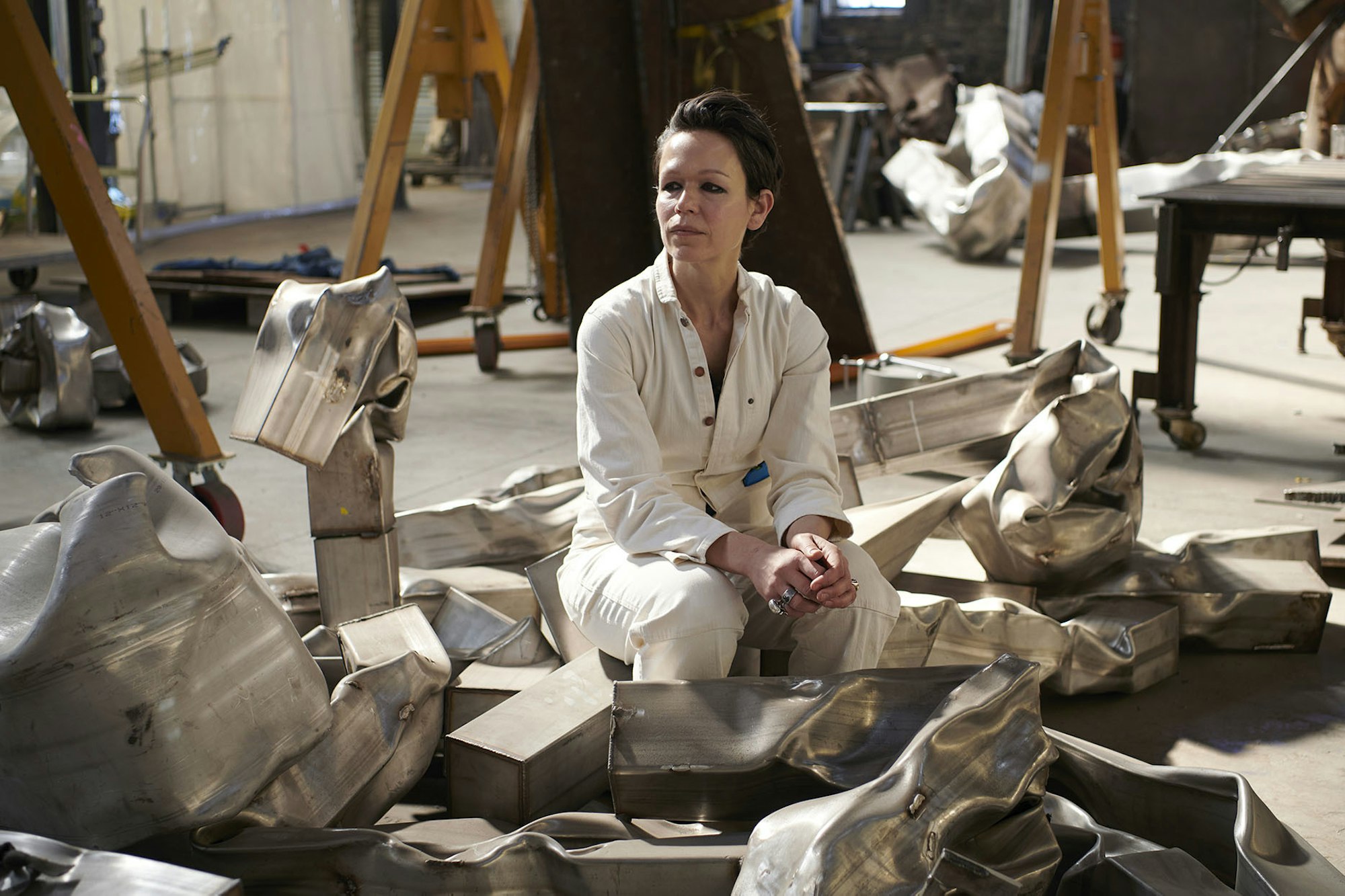 Carol Bove portrett. Bove sitter i verksteded sitt omgitt av bente jernmaterialer.